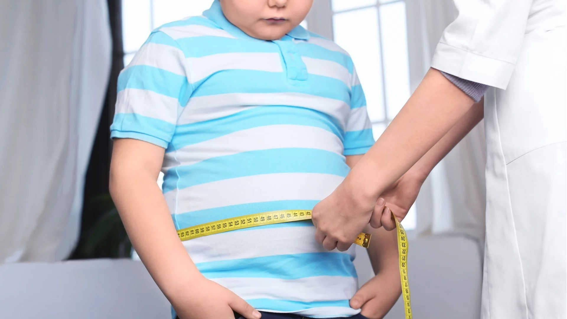 Fotografie zeigt ein Kind in blau-weiß gestreiftem T-Shirt, dessen Taillenumfang mit einem gelben Maßband von einer Person in weißem Kittel gemessen wird. Szene vermittelt medizinische Untersuchung oder Gesundheitscheck