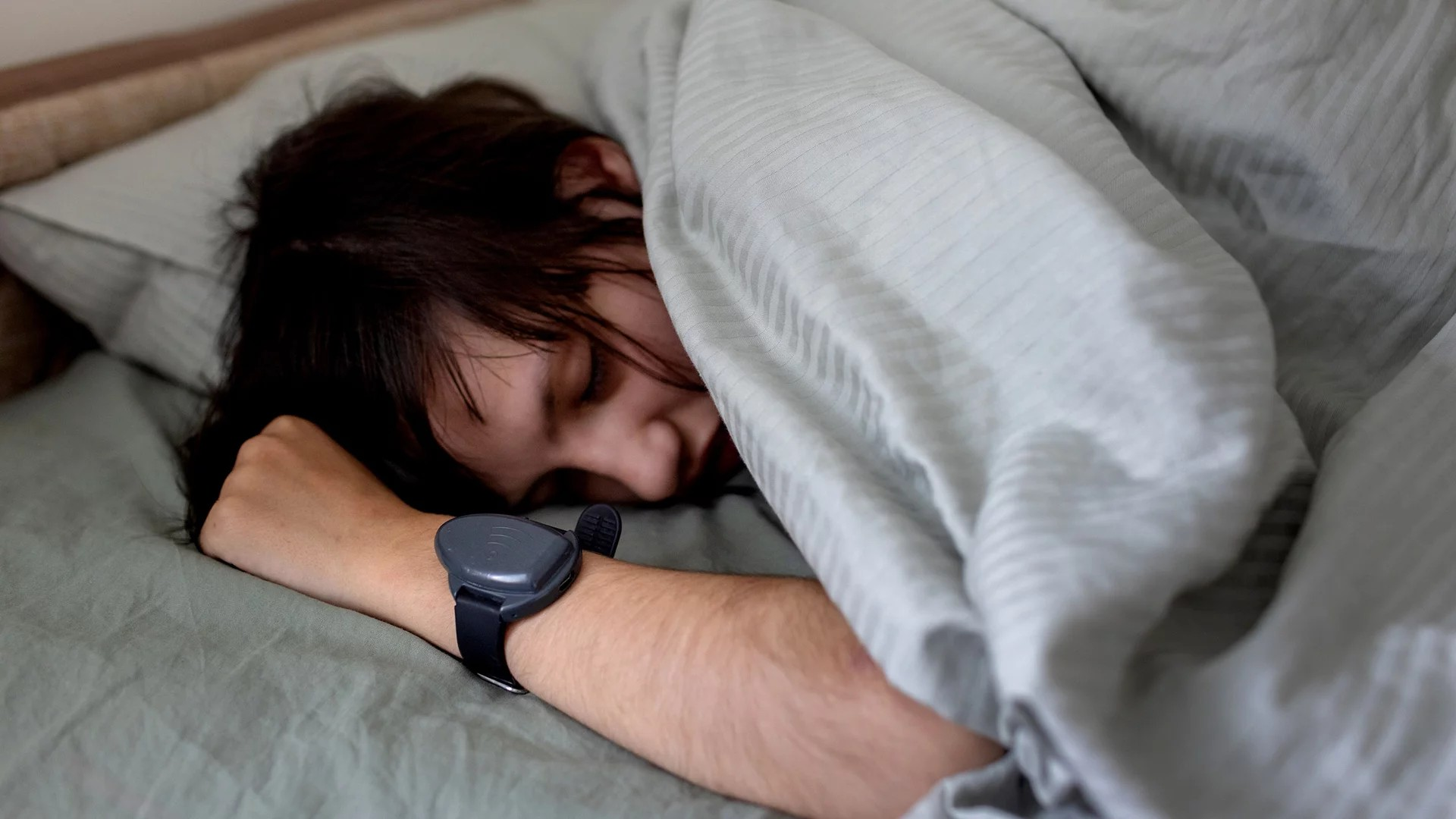 Fotografie einer schlafenden Person, die unter einer grauen Decke liegt und ein schwarzes Fitnessarmband am Handgelenk trägt. Fokus liegt auf entspanntem Gesichtsausdruck und ruhiger Schlafsituation in einem schlichten Schlafzimmer.