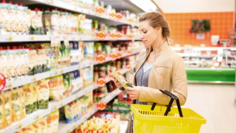 Fotografie einer Frau im Supermarkt, die ein Produkt aus einem Regal mit verschiedenen verpackten Lebensmitteln betrachtet. Sie hält einen gelben Einkaufskorb und steht vor Regalen mit bunten Verpackungen und Preisschildern.